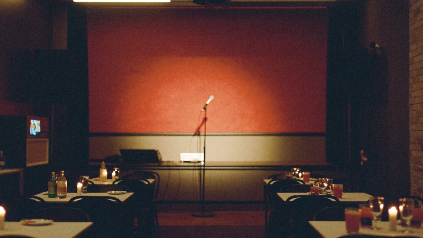 A small comedy club interior with cabaret tables and a single spotlit microphone stand on a low stage, warm red back wall, seen before doors open