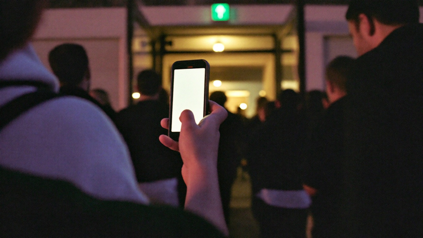 A hand holding up a glowing phone with a blank ticket screen in an entry line at night, a crowd seen from behind funneling through a warm-lit venue doorway, a green exit sign above the door