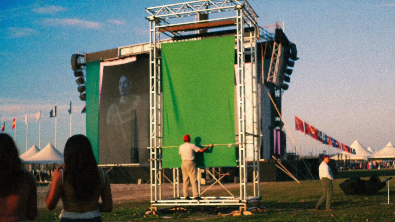 A festival crew member on a scaffold rig hanging a large plain green banner panel beside a stage at an outdoor event site, with flags and tents across the grounds in golden evening light