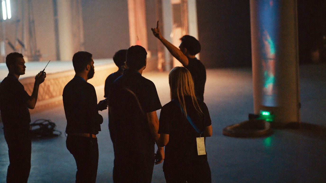 A pre-doors staff briefing in an empty venue: a small crew in lanyards holding two-way radios gathered near the stage under house lights before the crowd arrives, documentary style