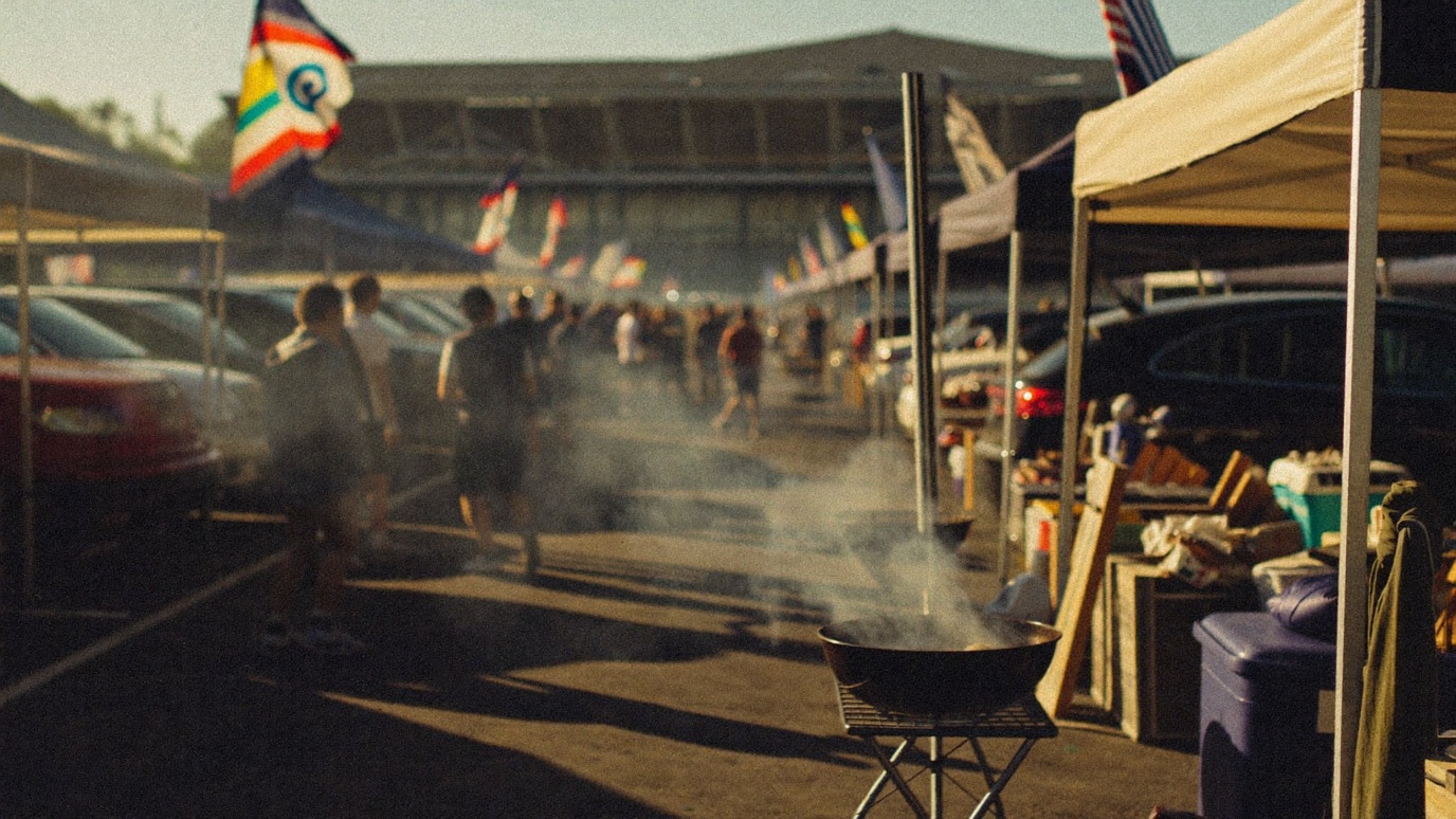 A stadium parking-lot tailgate at golden hour, a charcoal grill sending up smoke in the foreground beside pop-up tents and open car trunks, team flags along an aisle of tailgaters seen from behind, a large stadium grandstand out of focus on the horizon