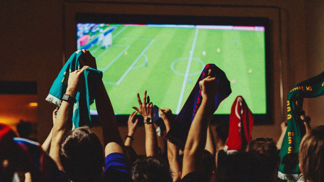A packed sports bar crowd seen from behind with arms and colorful scarves raised toward a large bright screen showing a blurred green soccer pitch, warm amber bar light mixed with the cool glow of the screen
