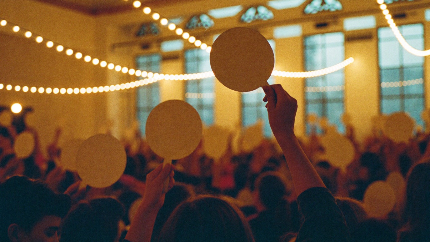 A crowd seen from behind at a charity benefit, arms raised holding plain blank round auction paddles in a warm-lit ballroom with strings of cafe lights and tall blue-lit windows behind them