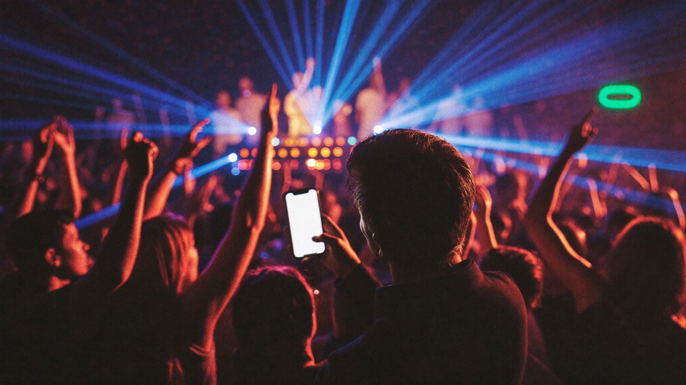 A dense club dancefloor seen from behind the DJ booth, hands raised toward stage lasers cutting through haze, a promoter at the edge of the crowd glancing at a glowing phone, deep magenta and blue club light