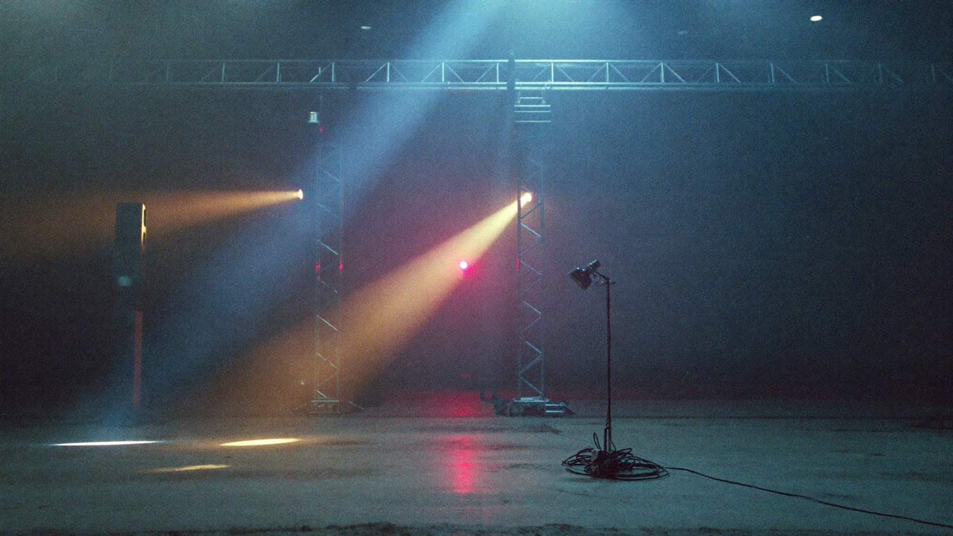 A raw industrial warehouse before doors, trussing and moving-head lights being rigged over an empty concrete floor, atmospheric haze catching cool blue and warm amber beams, a single work lamp lighting a coiled power run in the foreground