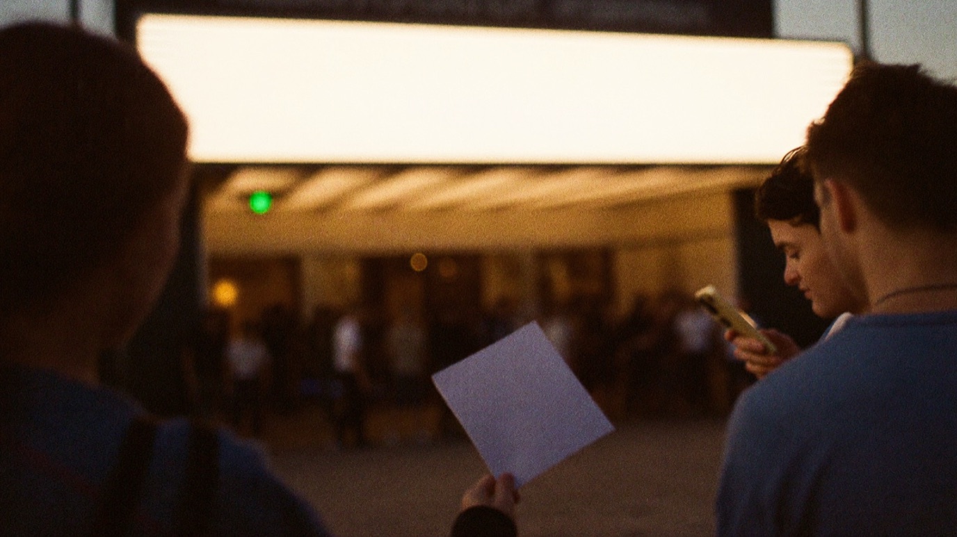 Two silhouetted promoters in the foreground outside a small venue at dusk, one holding up a blank pale flyer while a companion checks a phone, with a blank glowing marquee lightbox and a blurred line of people at the lit entrance behind them, warm golden light against blue shadow and a small green exit light
