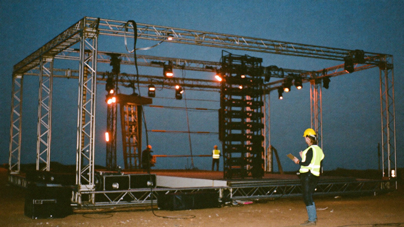 A production manager in a hard hat holding a clipboard in front of a half-built festival main stage at dusk, scaffolding truss and stage crew working under work lights