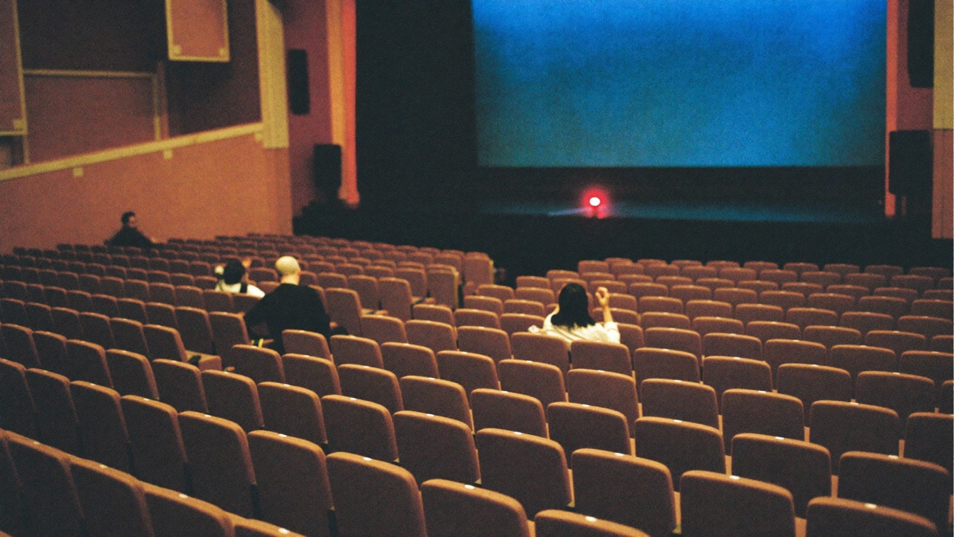 A large theater with rows of mostly empty seats and only a few scattered people facing a dim stage, a small red light in the distance, conveying an event that did not sell